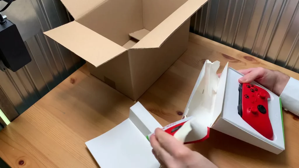 Auspacken des Gamecontrollers Roter Gameontroller wird aus seiner Verkaufsverpackung genommen, neben einem leeren Pappkarton